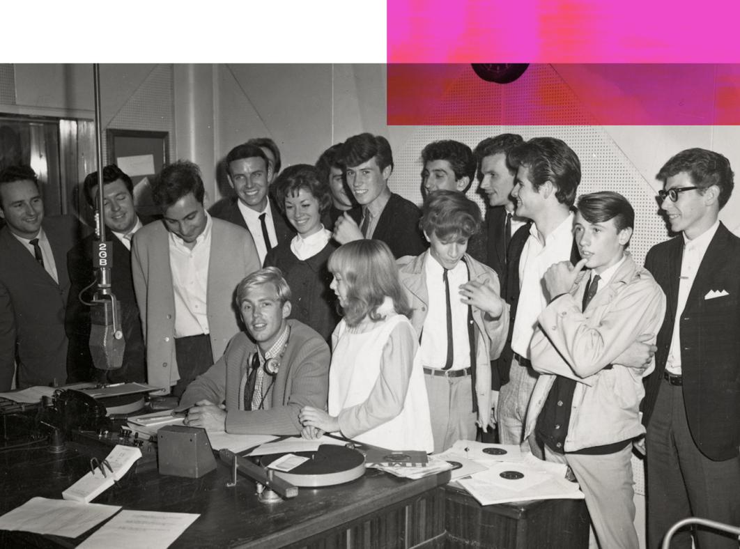 A man seated at a desk in a radio studio surrounded by young people.