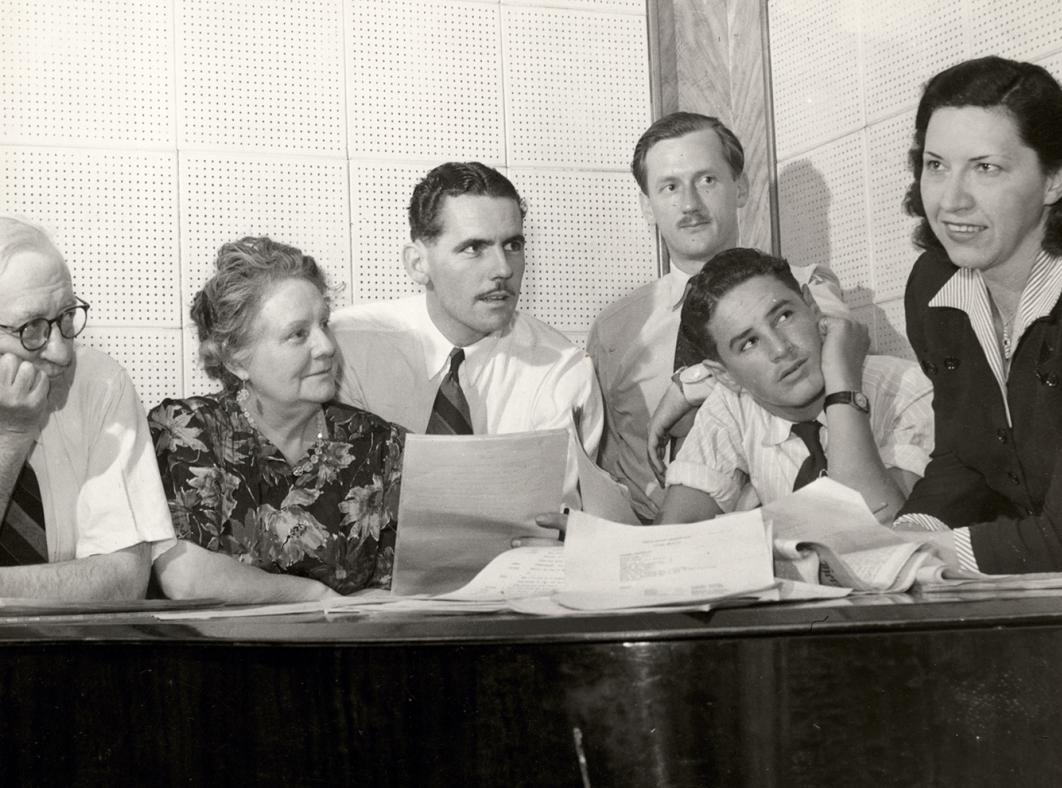 A group of people sitting at a desk holding papers and looking sideways at radio executive Grace Gibson