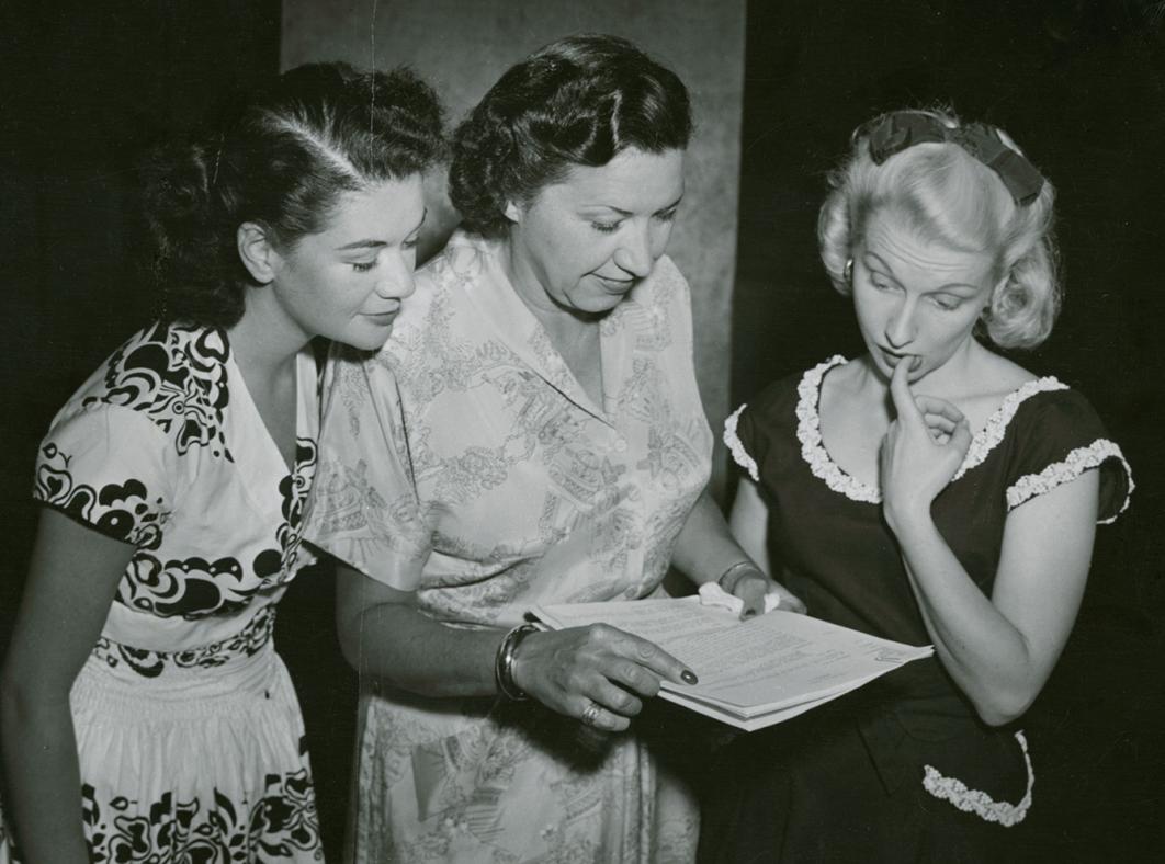 Radio personalities Betty McDowall and Madi Hedd standing either side of Grace Gibson. They are all looking at some papers held by Gibson.