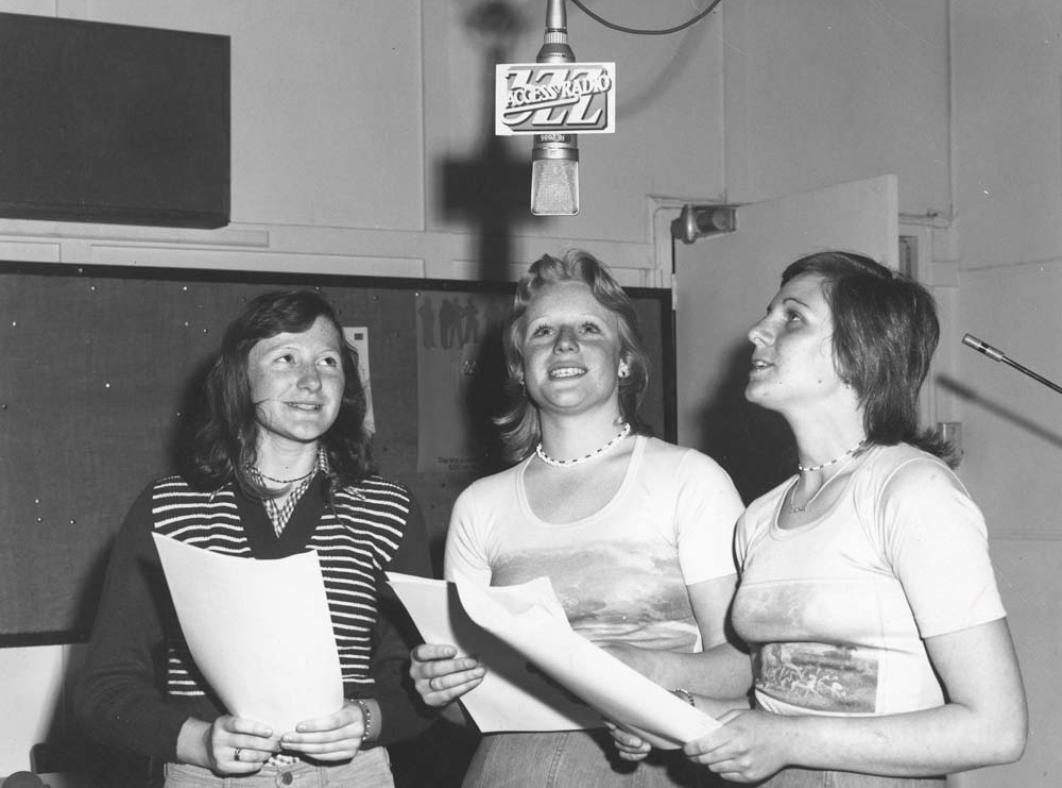 Three women holding scripts in a radio studio are standing around a microphone suspended above them