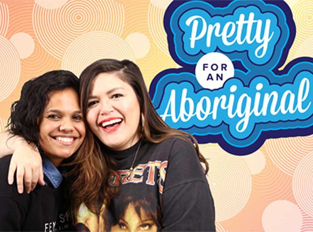 Two women, Miranda Tapsell and Nakkiah Lui, embracing each other and smiling. They are pictured in front of a sign saying Pretty for an Aboriginal.