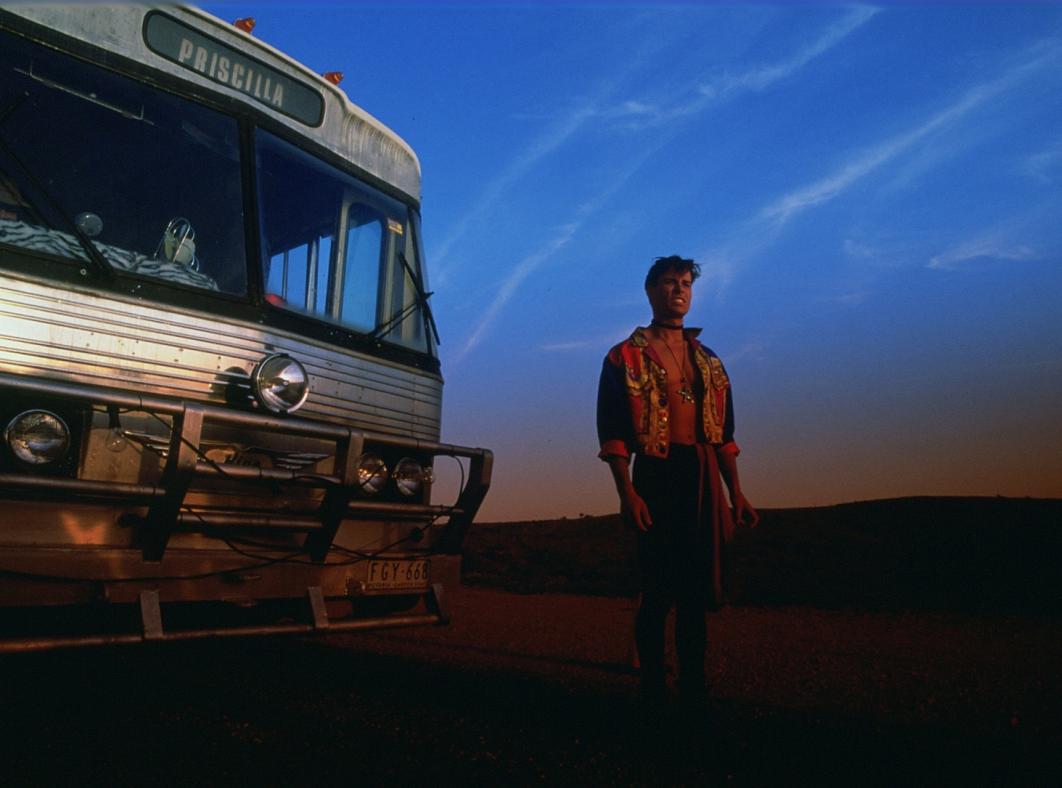 Guy Pearce standing in front of a bus with Priscilla written above the window. The sky is dark with an orange glow on the distant horizon