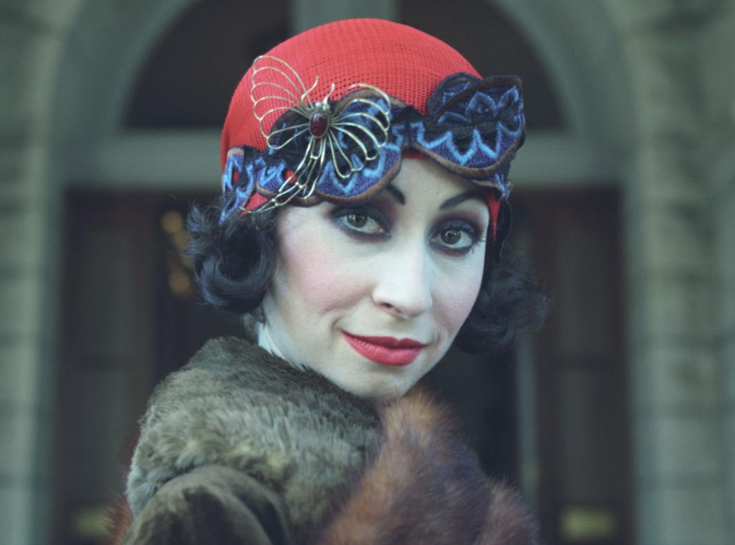 Close up of a woman in a 1920s style hat and coat looking sideways at the camera.