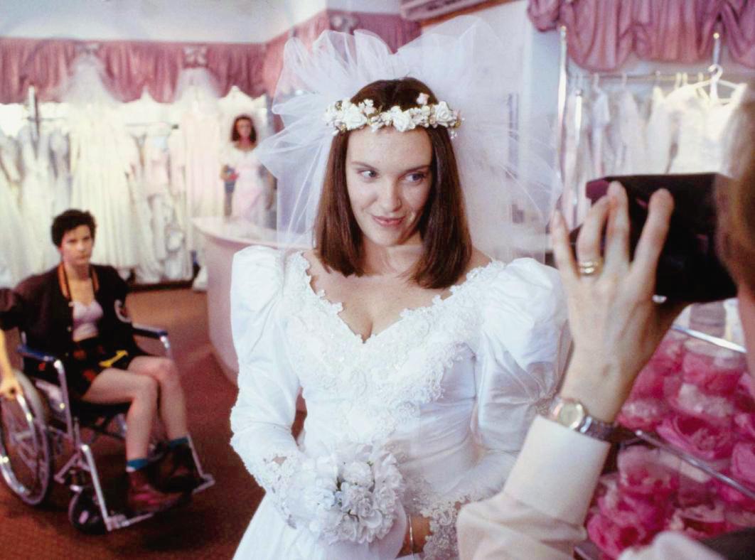 A young woman in a bridal store wearing a wedding dress and another woman in a wheelchair behind her in a scene from Muriel's Wedding.
