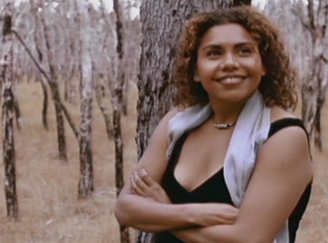 Waist-up shot of actor Deborah Mailman leaning on a tree with her arms folded and smiling.