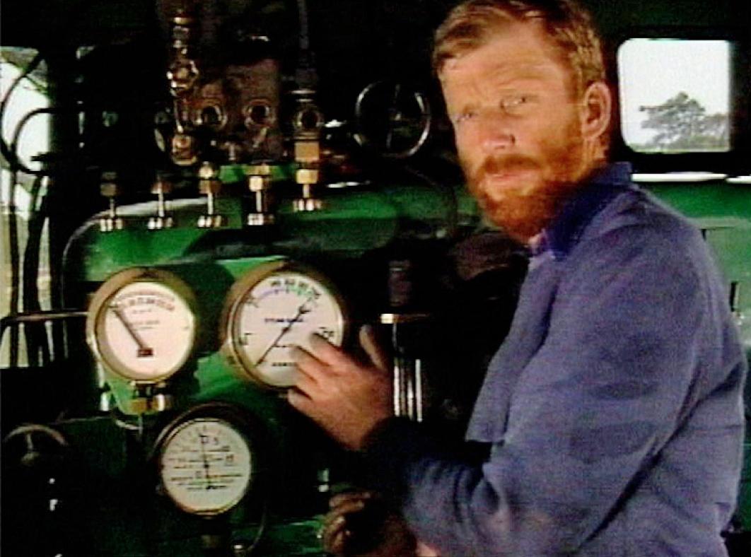 A train engineer stands at the controls of a steam engine. He is pointing to a gauge and facing towards the camera. 