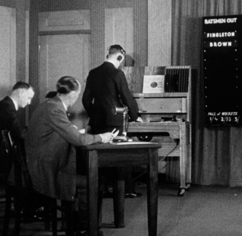 people in a radio studio with a cricket scoreboard on the wall in front of them
