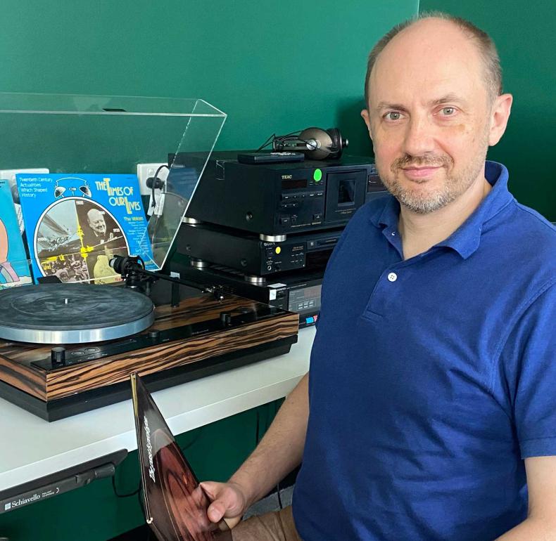 NFSA curator Simon Smith, seated and looking up at the camera. He is sitting at a desk with a turntable and vinyl records on it.