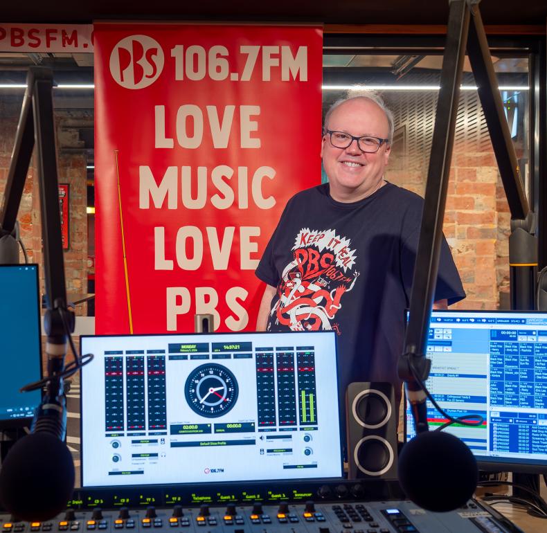 A man stands behind a radio station mixing desk and monitor smiling. He stands in front of a banner that says "106.7FM Love Music Love PBS".
