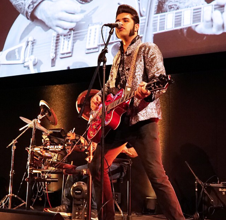 Elvis tribute singer Jerikye Williams performing on stage playing a guitar and standing in front of a drummer with a cinema screen behind them