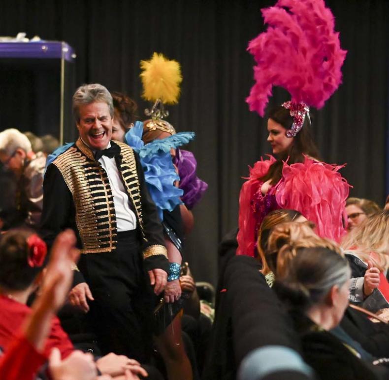 Two ballroom dancers standing next to the audience in a crowded cinema after a performance. They are both laughing. The male dancer wears a black and gold vest with a bow tie while the female dancer wears a pink costume with a high pink-feathered headdress