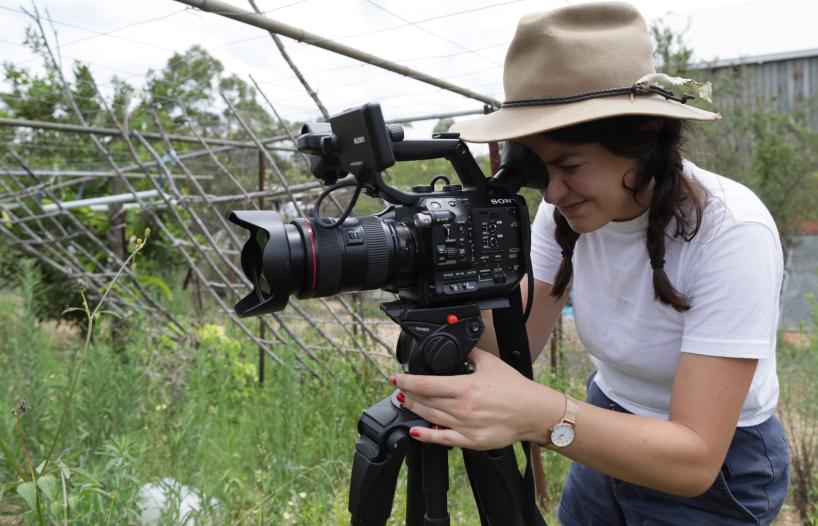 A young woman with her head tilted forward looking into the viewer of a movie camera on a tripid.