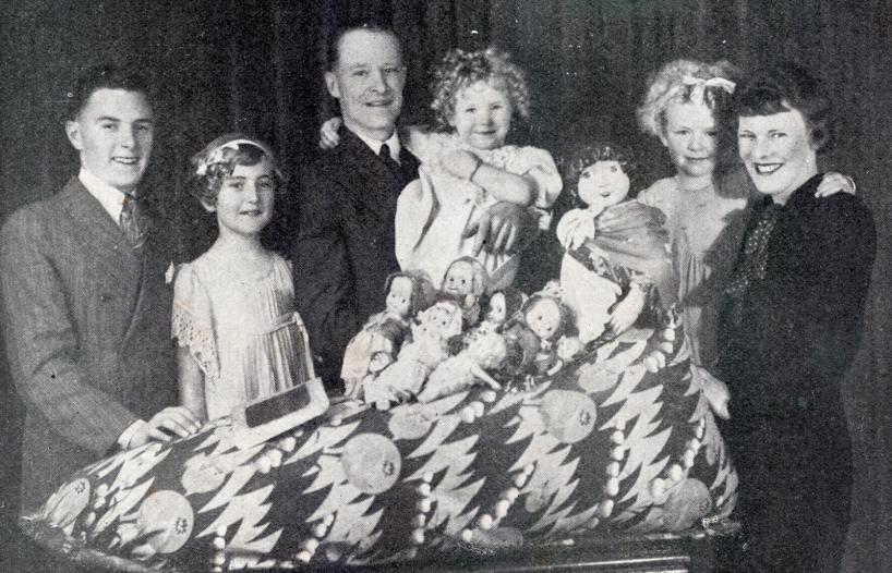 Two men and a woman posing with three kids and some dolls. They are all smiling at the camera. 