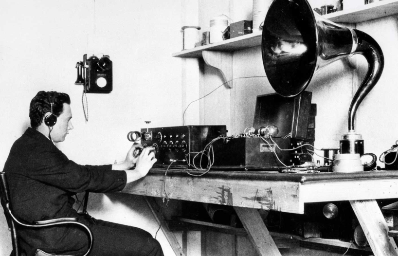 A man sitting at a desk in a 1920s radio studio with radio equipment and a gramophone next to him