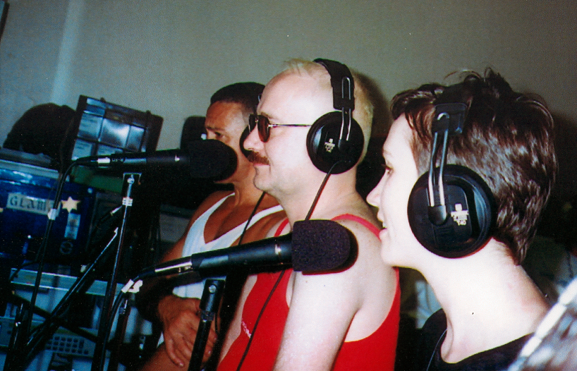 Three people pictured in profile, wearing headphones and standing side by side in front of microphones in a radio studio.