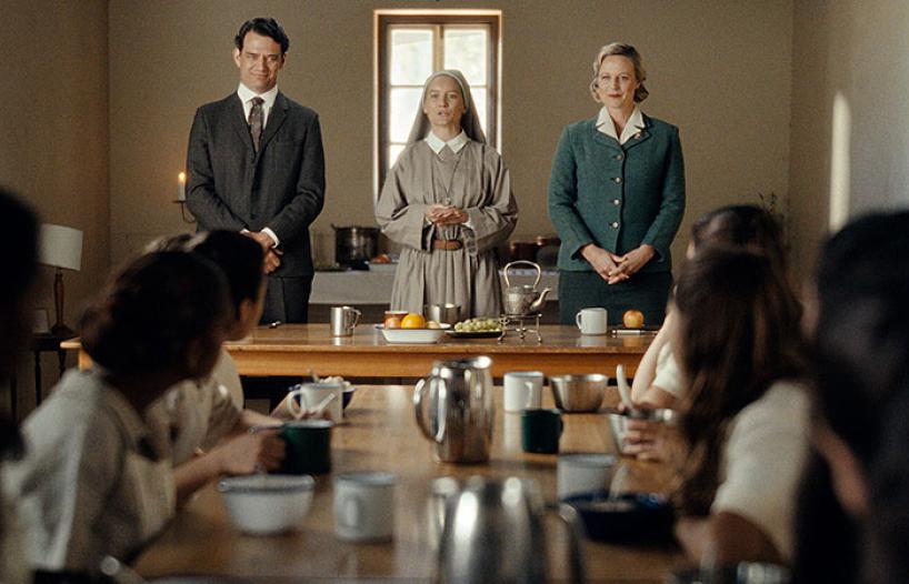 A man and a woman standing either side of a nun, addressing a room full of young First Nations schoolgirls.