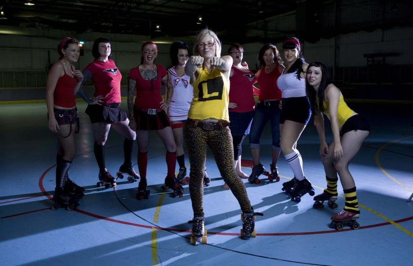 Nine female roller derby players wearing roller skates and standing in a semi-circle, posing. The middle player stands in front of the others and has her arms outstretched and hands bunched into fists.