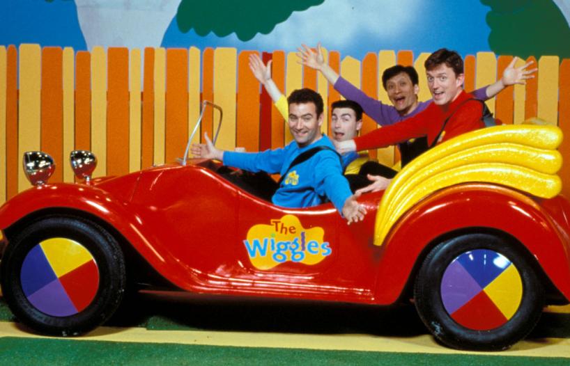 The four members of children's music group the Wiggles, seated in a lifesized, open-topped red toy car with the Wiggles logo on the side door. The car is parked next to a colourful fence on a studio set and the Wiggles are all extending their arms and smiling.