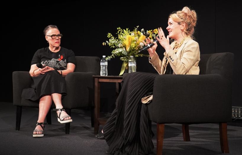 Dr Brenda Croft interviewing filmmaker Jazz Money on stage at Arc Cinema at the NFSA. Jazz is speaking into a microphone and there is a vase of native yellow flowers on a table between them.