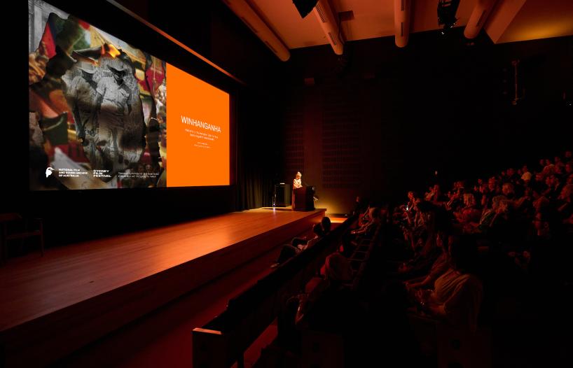 A view of the cinema audience from the side at a screening of WINHANGANHA. Director Jazz Money stands behind a lectern in front of the screen showing an image from the film