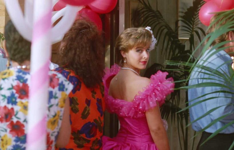 A young woman in a frilly hot pink bridesmaid dress walking away from the camera and looking back over her shoulder in a scene from Muriel's Wedding