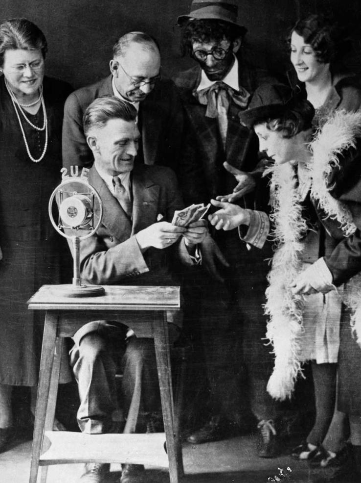 A group of people standing around a man seated at a small table behind a 2UE microphone