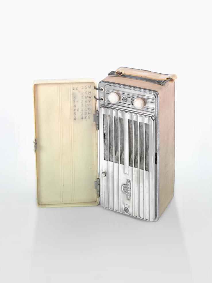 Portable radio from the 1940s. It is a tall, rectangular box shape with a front cover and handle on top. Inside there are dials and a speaker.