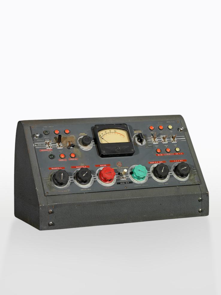 A mixing console for a radio broadcast studio. It is a triangular, wedge shape in a grey metal housing with a lot of differently coloured dials, switches and knobs on the front.