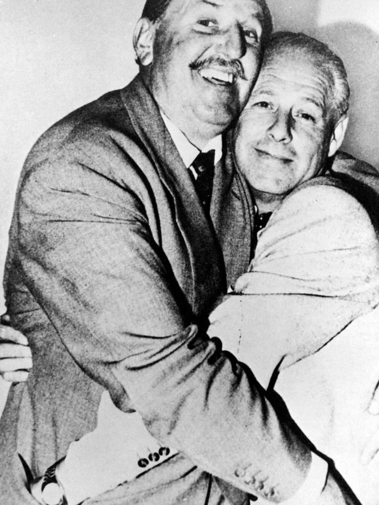 Two middle-aged men embracing and smiling. They are looking at the camera and have their hands in each other's pockets.