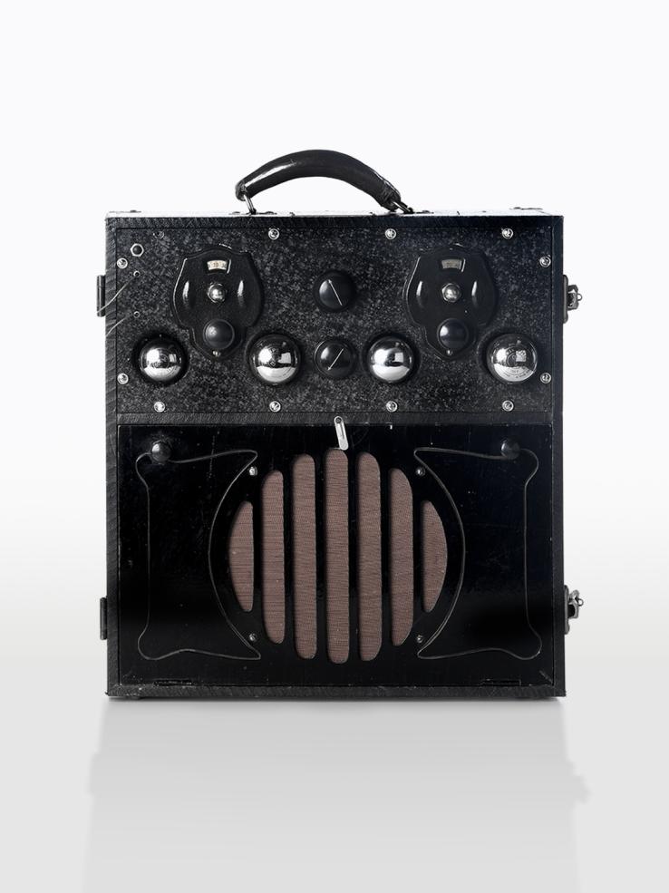 A radio received in a black box with round speaker and dials on the front.  It has a carry handle on the top.