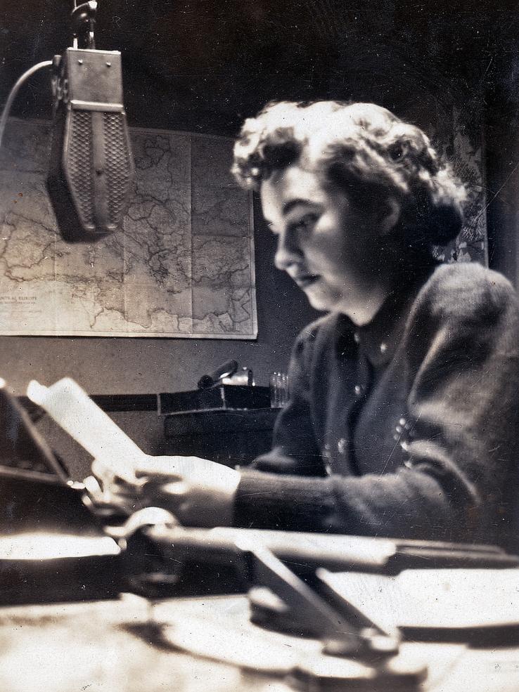 A woman sitting at a desk, side on to the camera. She is holding papers and has a radio microphone in front of her.