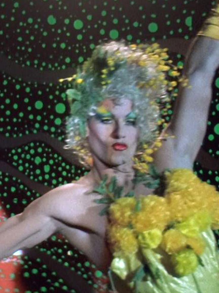 Guy Pearce on stage wearing a blond wig and a yellow dress with yellow pom poms that resemble wattle flowers. His lips are pursed and his arms stretched out to the sides