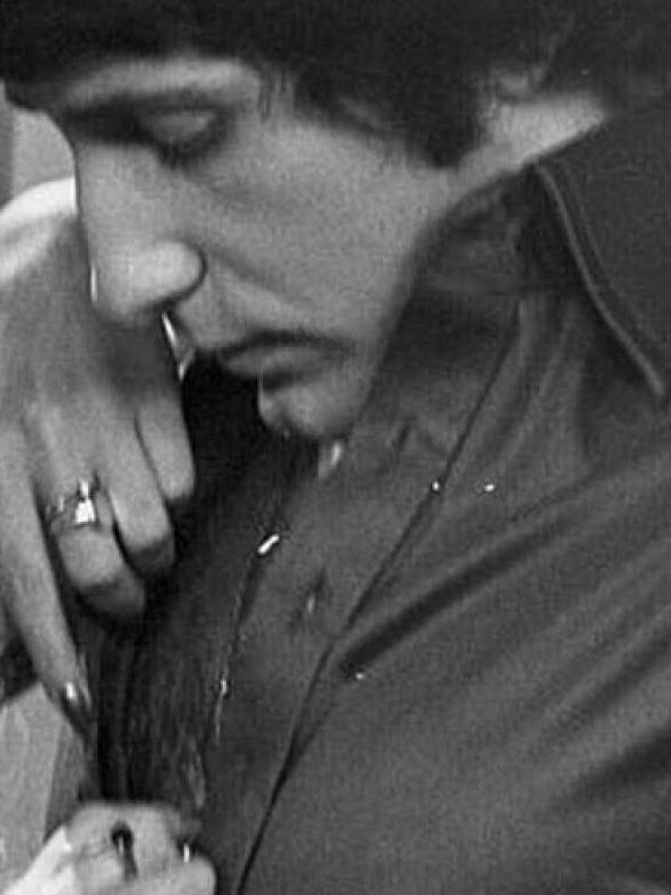 A man wearing a short with a wide collar looks down as a a woman's hands are undoing his shirt buttons