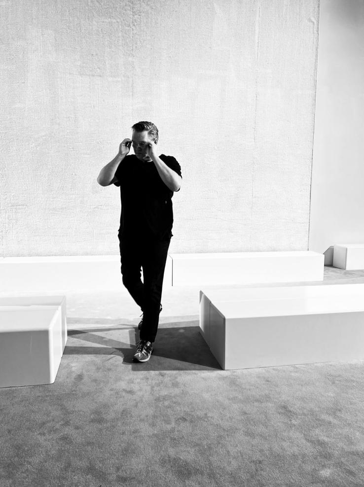 Artist Gary Sinclair walking through a gallery exhibition space, wearing black shirt and jeans and adjusting his glasses