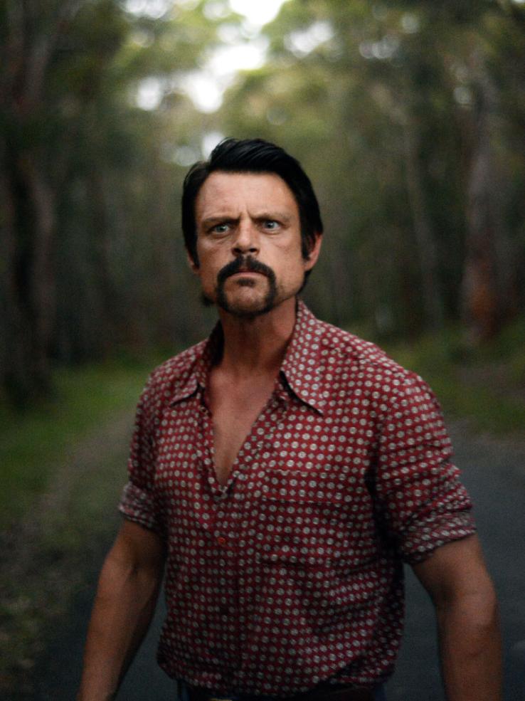 upper body shot of a menacing looking man standing in a road near a forest.