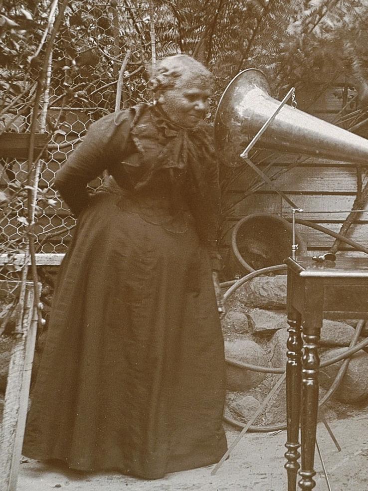 First Nations woman Fanny Cochrane Smith wearing a dark dress and leaning into the horn of a gramophone from the 1890s
