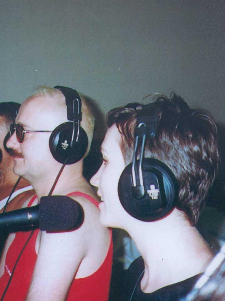 Two people wearing headphones and standing behind microphones in a radio studio.