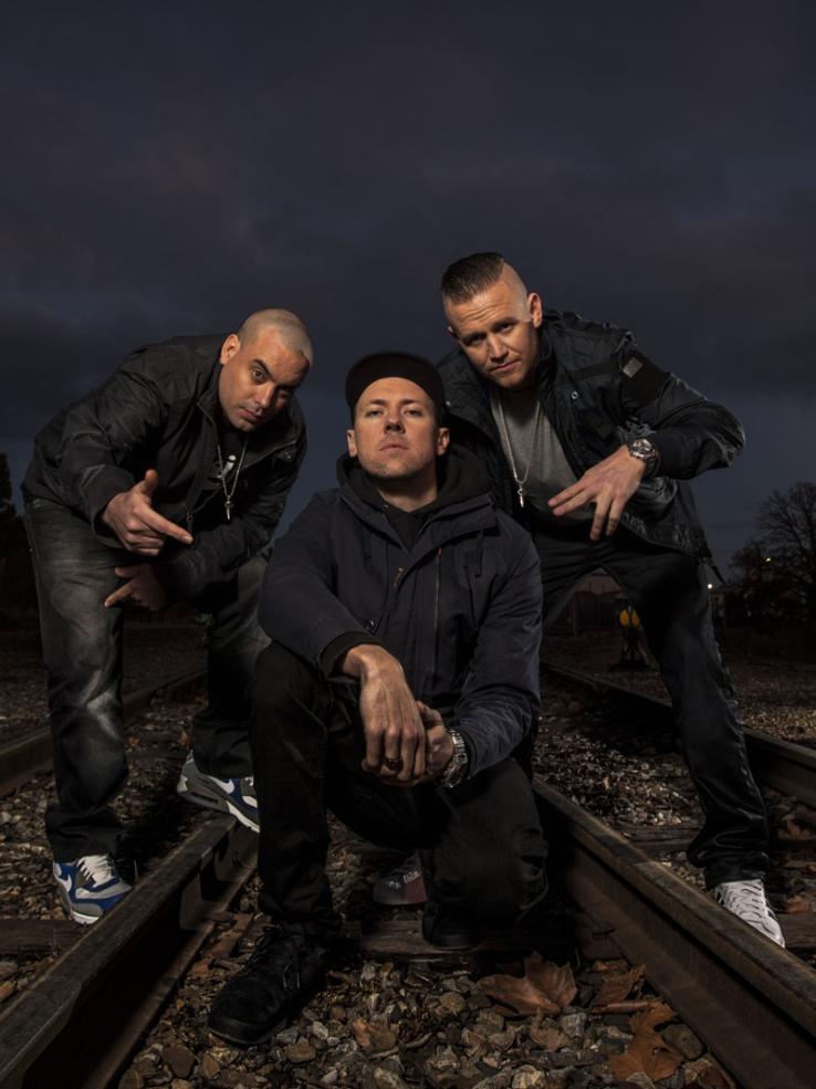 Three members of hip hop group Hilltop Hoods wearing dark clothing and posing outside on railway tracks at night.