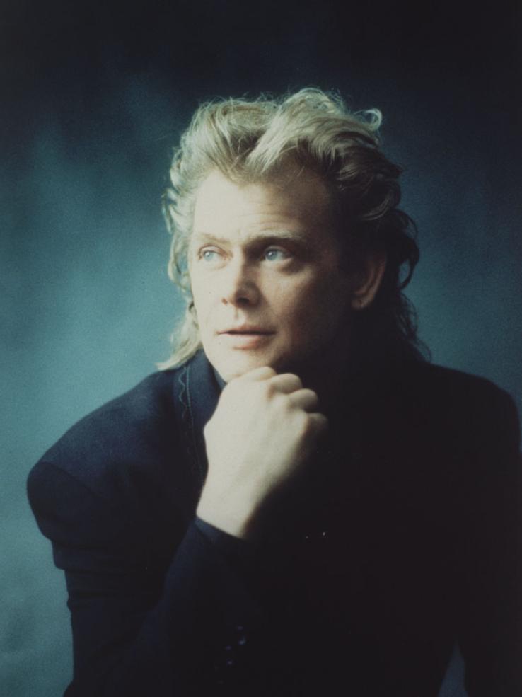 John Farnham wearing a dark jacket and looking away from the camera, with his chin resting in his hand.