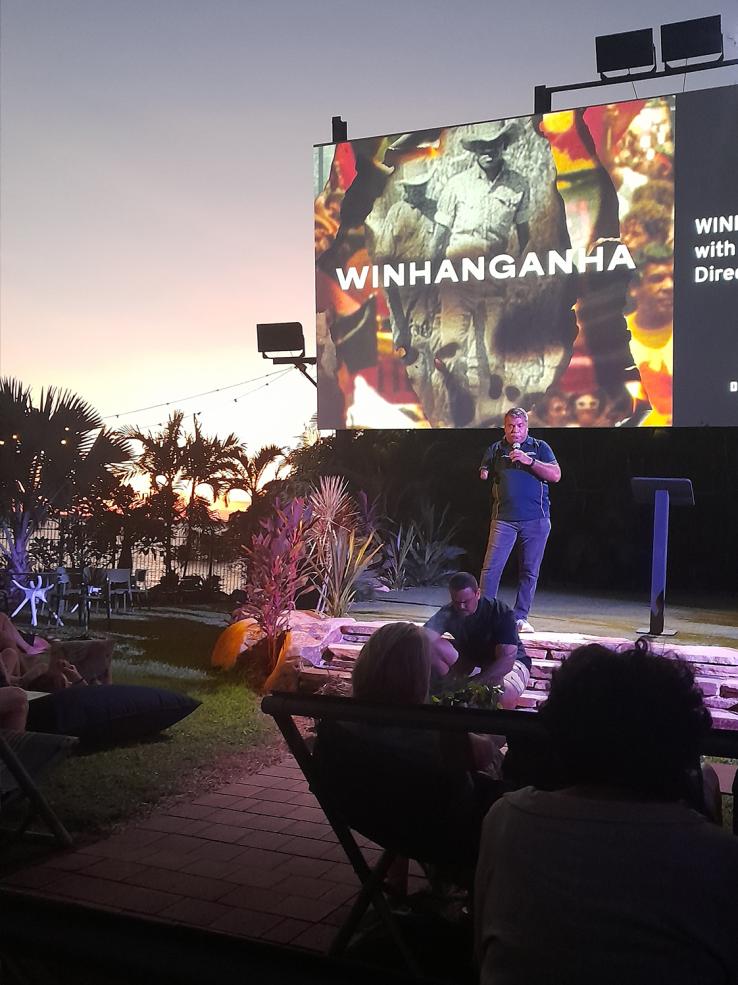 A man on stage introduces an outdoor cinema screening of the film WINHANGANHA