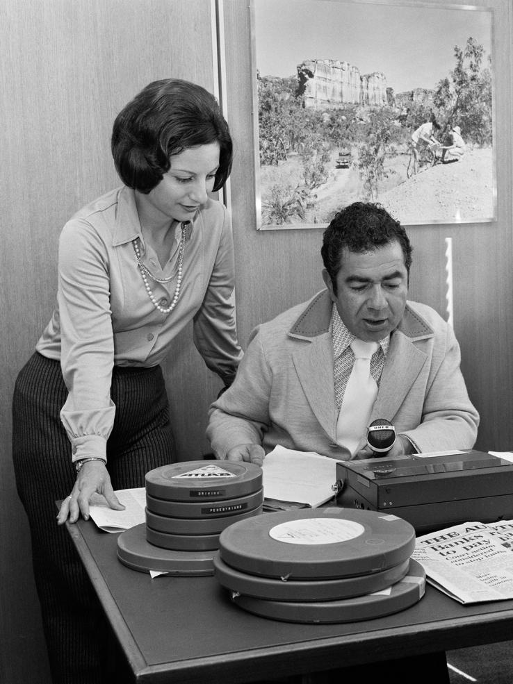 Radio presenter Sophia Catharios stands behind fellow presenter Takis Kaldis, who is seated behind a desk. Takis holds a microphone and is speaking into a recorder. On the desk are several circular tape canisters.