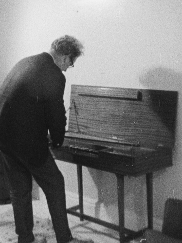 Composer Ron Grainer standing hunched over playing a small upright piano.