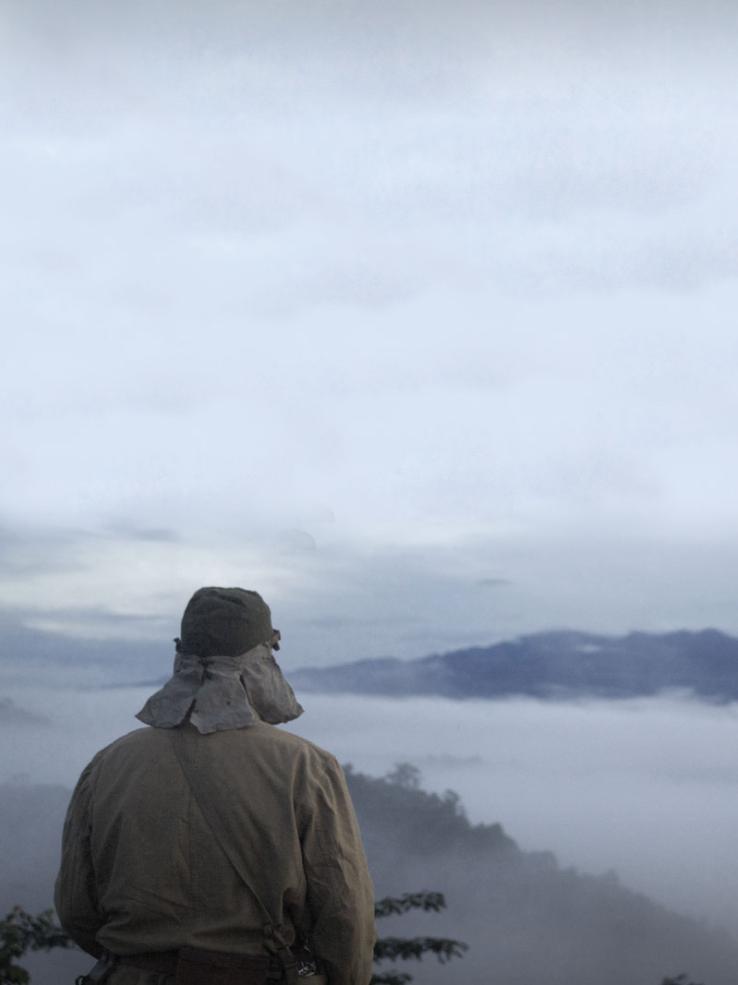 A Japanese soldier sits on a hilltop looking out on a misty mountainous landscape.