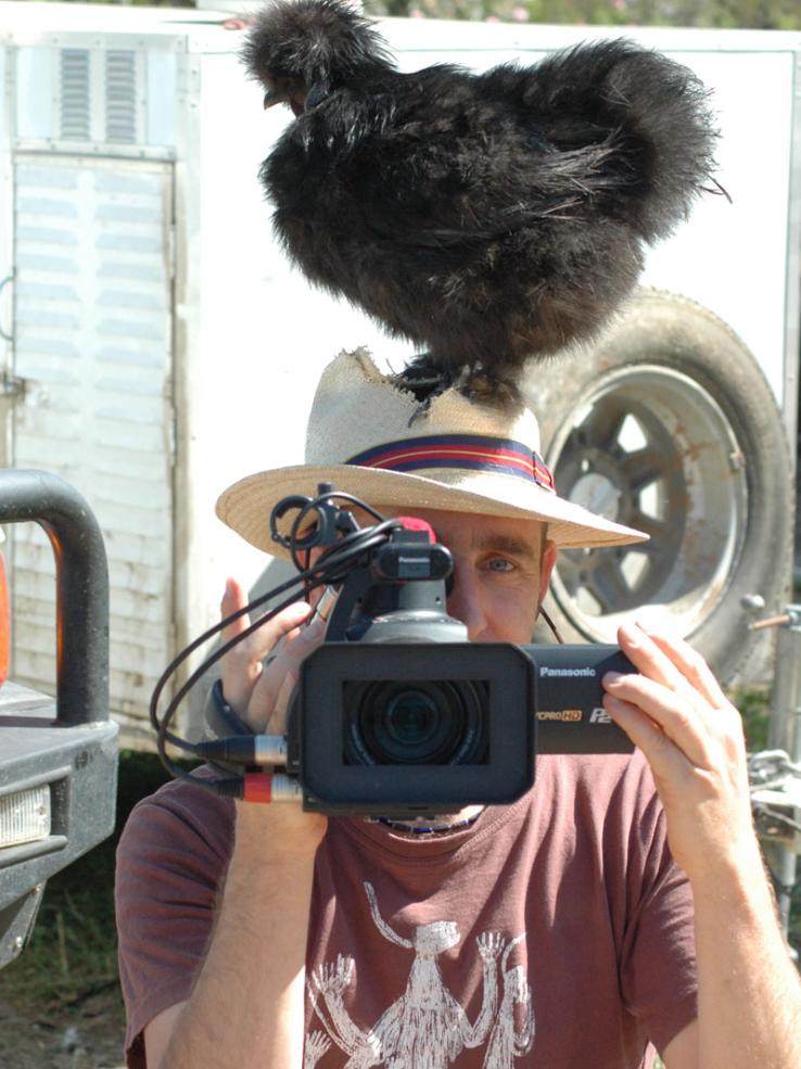 A man points a video camera while a black fluffy chicken perches on top of the panama hat he is wearing.