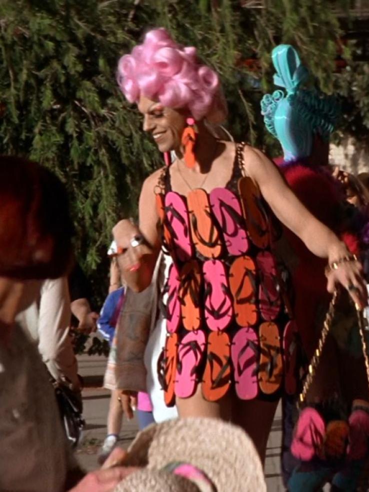 Hugo Weaving with a bright pink wig and wearing a dress made out of pink and orange flip-flops. He is walking down a footpath in drag make-up and attracting surprised looks from passersby
