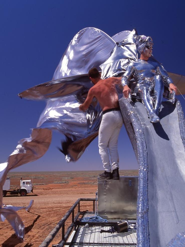 Guy Pearce sitting on a giant silver stilleto shoe on top of the Priscilla bus.