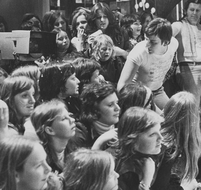 A crowd of teenagers in a TV studio sitting behind the cameraman.