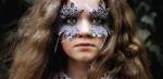A close up of a young girl looking directly at the camera. She is wearing feather decorations around her eyes.