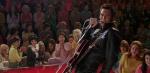 Austin Butler as Elvis Presley singing to a TV audience of adoring female fans in the movie ELVIS. He has a guitar strapped to him and is wearing a black leather suit.