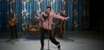 Austin Butler as Elvis Presely singing with his 4-piece band on stage in a recreation of a 1950s TV studio. Elvis is wearing a pink blazer and the group are performing in front of a turqouise curtain with sun symbols on it